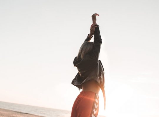 A fashionable woman stands with arms raised on a beach at sunset, exuding style and freedom.